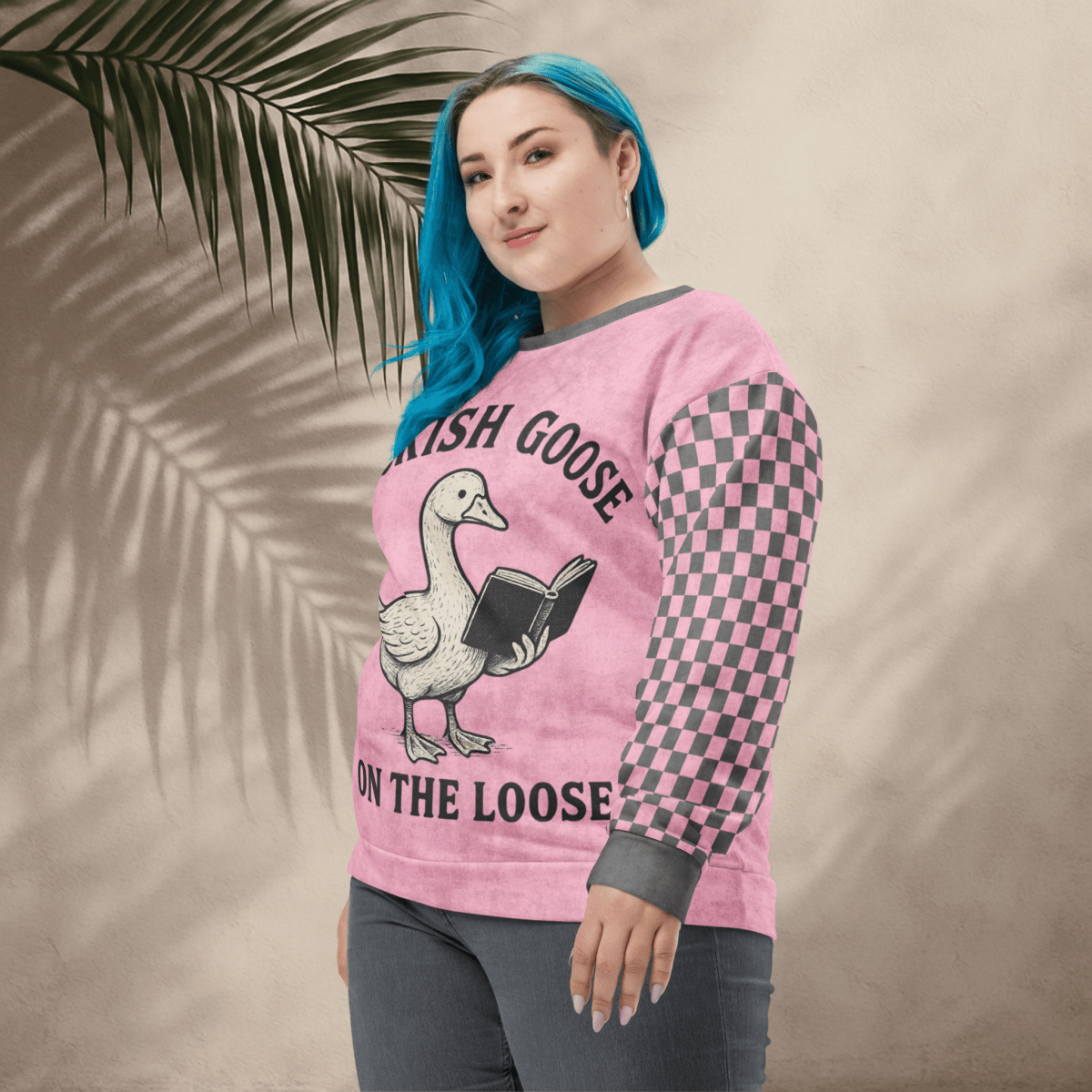 Person wearing a pink sweater with a graphic design and text, standing against a beige wall with a leafy branch.