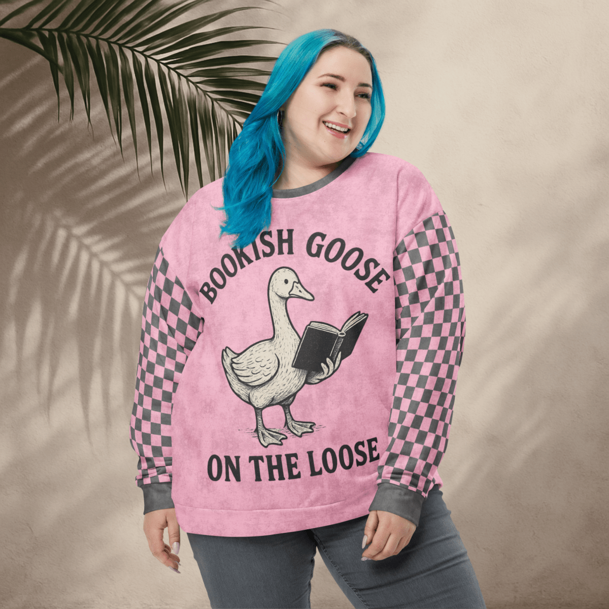 Person wearing a pink sweater with a goose graphic and text, standing against a beige wall with a leafy branch.