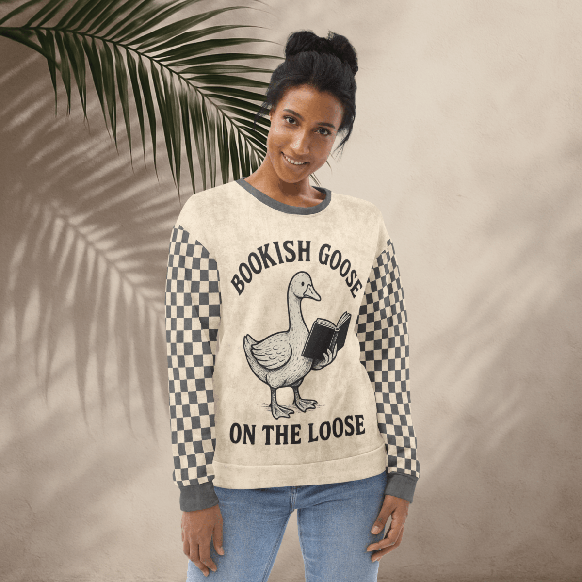 Woman wearing a sweater with a goose graphic and text, standing against a beige wall with a leafy branch.