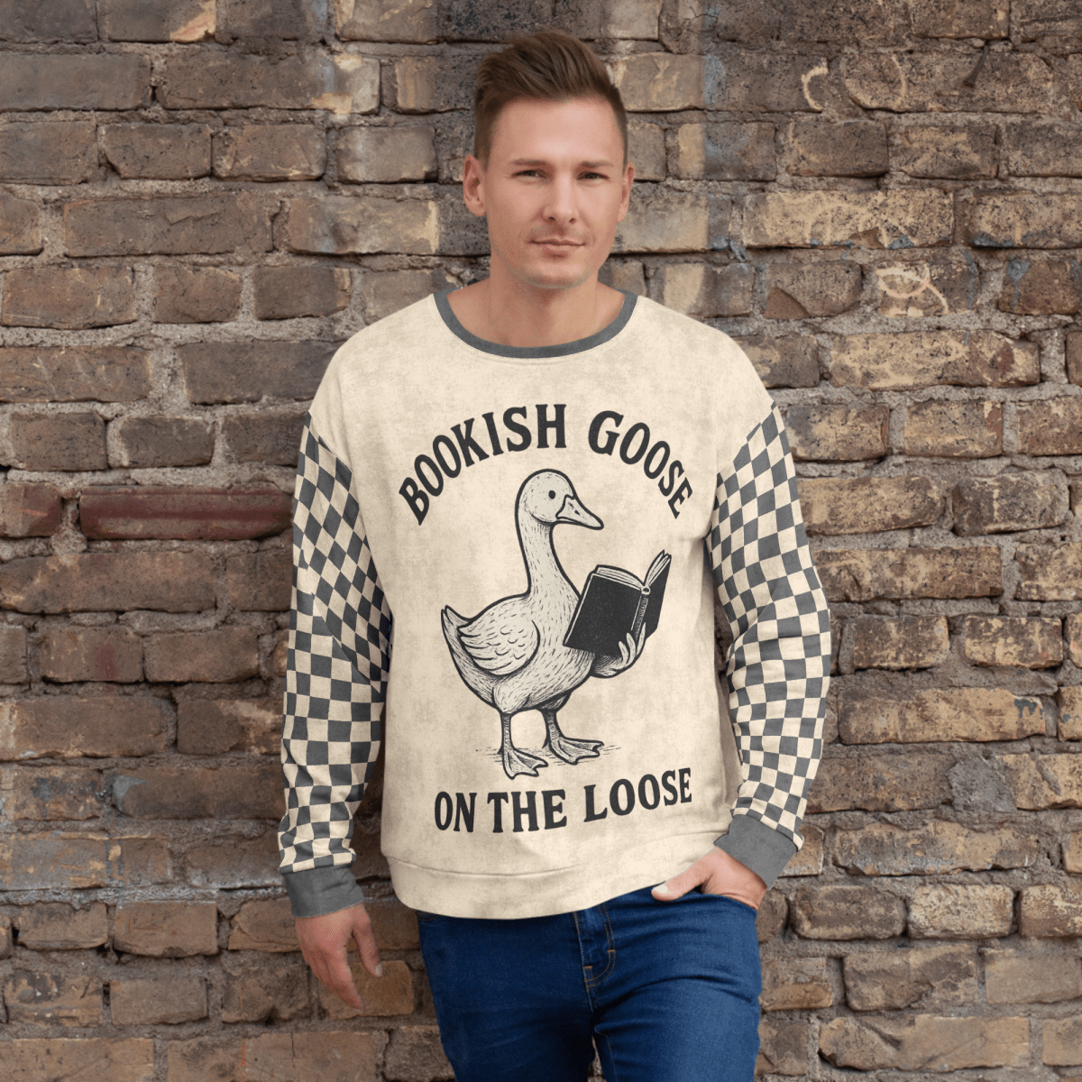 Man wearing a sweater with a graphic of a goose holding a book and text 'Bookish Goose on the Loose' against a stone wall.