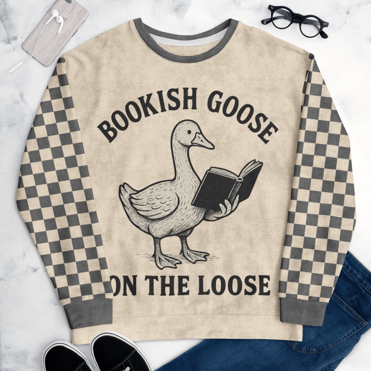 Beige sweater with a goose holding a book and text 'Bookish Goose on the Loose' on a marble surface.
