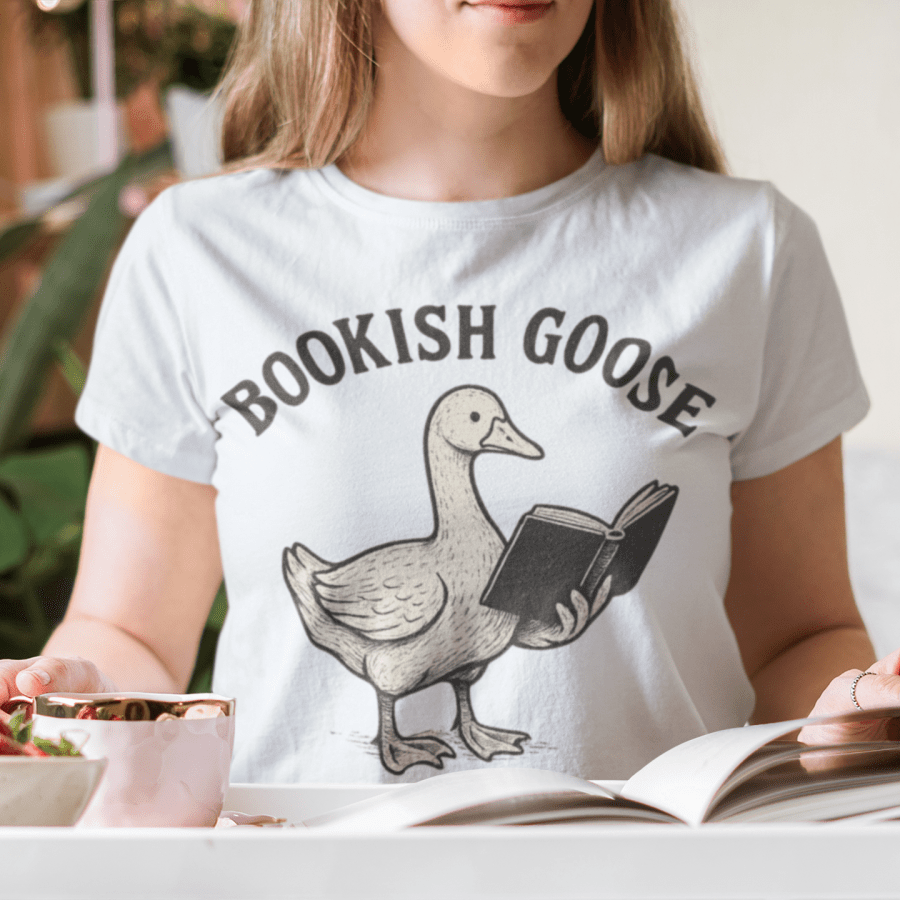 Person wearing a white t-shirt with 'Bookish Goose' design, holding an open book and strawberries.