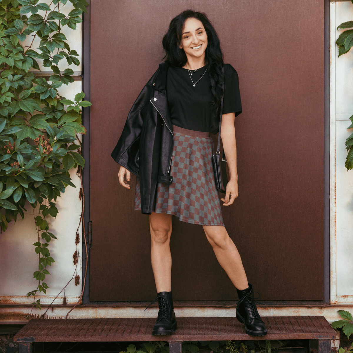Woman wearing burgundy and gray checkered skirt outdoors with a brown door and greenery