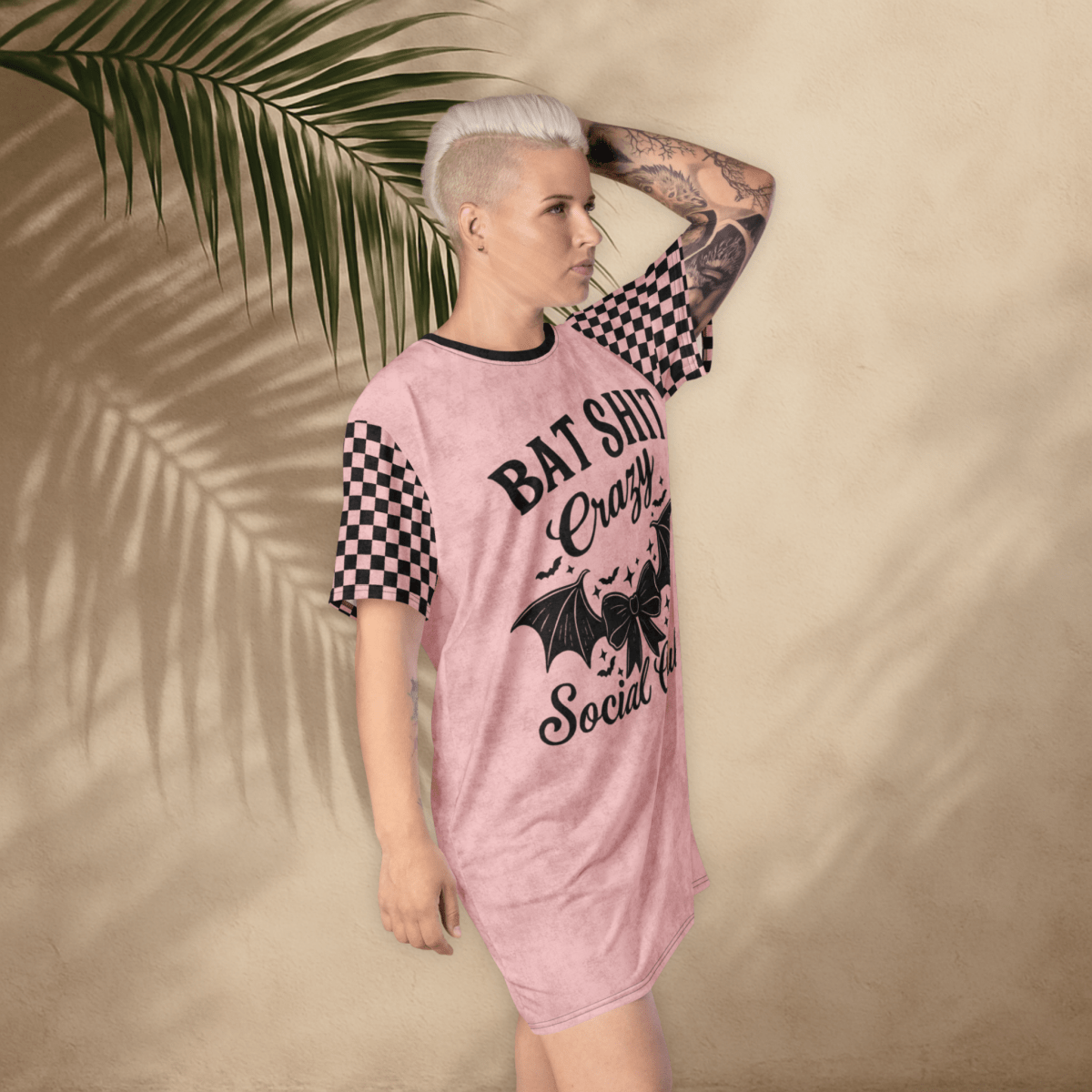 A person with platinum-blonde hair and arm tattoos poses in a pink t-shirt dress against a tan wall with palm leaf shadows. The dress features black checkerboard sleeves and a bold black graphic that reads "BAT SHIT Crazy Social Club" with a bat-winged bow and stars. The person stands with one hand on their head, tilting slightly forward.