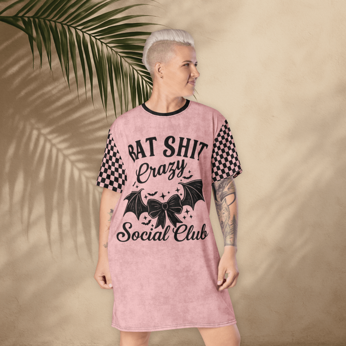 A person with platinum-blonde short hair and visible arm tattoos stands in front of a tan stucco wall with palm leaf shadows, wearing a pink t-shirt dress. The dress features black checkerboard sleeves and bold black text on the front that reads "BAT SHIT Crazy Social Club," accompanied by graphic elements like a bow, bat wings, and scattered stars. The person faces forward with a slight smile and hands relaxed at their sides.