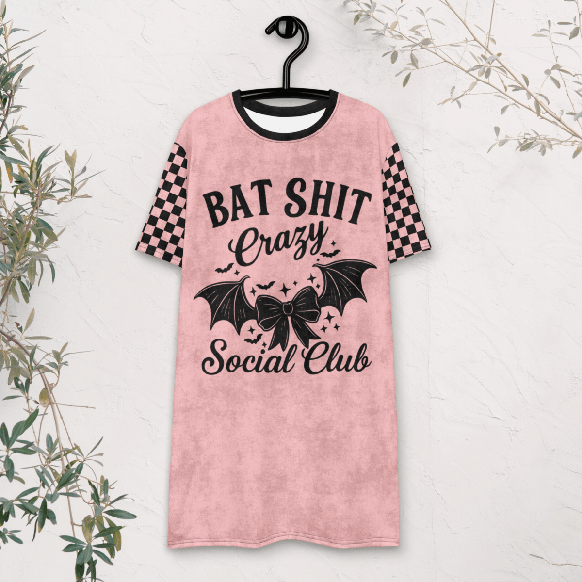 A pink t-shirt dress is displayed on a black hanger against a white textured wall. The dress features black checkerboard short sleeves and a large front graphic in black that says "BAT SHIT Crazy Social Club," surrounded by bats, stars, and a central bow. Some foliage partially frames the edge of the photo.