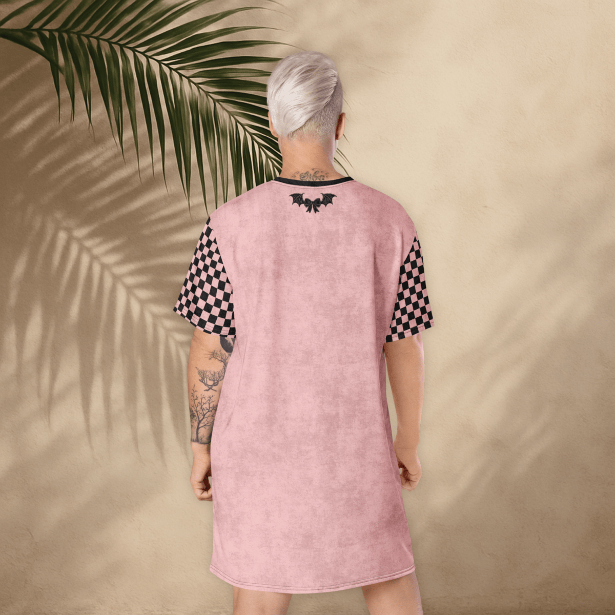 A person with platinum-blonde hair stands with their back to the camera, wearing a pink t-shirt dress. The dress has black checkerboard sleeves and a small black bat-and-bow graphic printed between the shoulder blades. The background is a light tan stucco wall with large palm leaf shadows cast across it. The person’s tattoos are visible on both arms.