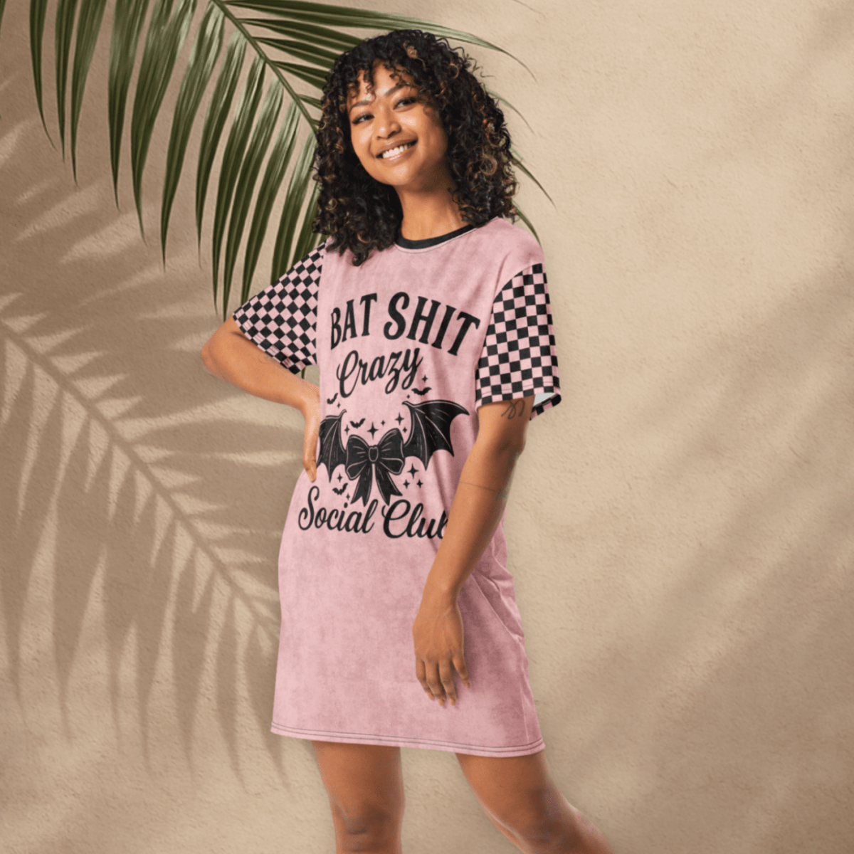 A person with medium-toned skin and curly dark hair poses smiling in a pink t-shirt dress in front of a tan stucco wall with palm leaf shadows. The dress has black checkerboard sleeves and a front design that reads "BAT SHIT Crazy Social Club" in bold black font, with a bow and bat wings below the text. The person stands on terracotta tiles and wears white platform sandals.