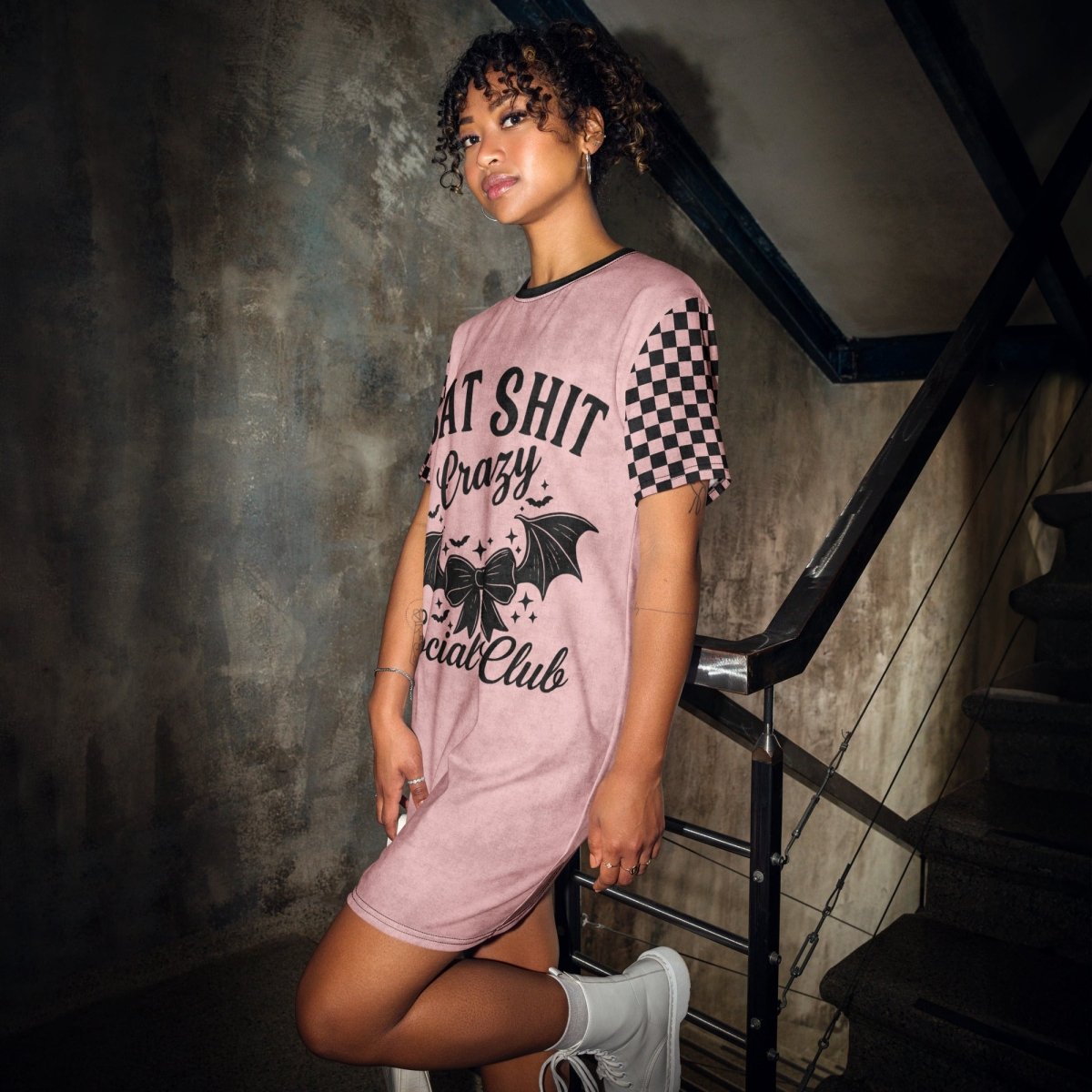 A young woman with curly hair stands on an industrial-style staircase in front of a weathered concrete wall. She wears a pink t-shirt dress with black checkerboard sleeves and a large black graphic on the front that says "BAT SHIT Crazy Social Club." The design includes a bow with bat wings, small stars, and bold gothic text. She pairs the dress with white lace-up boots and gazes confidently at the camera.