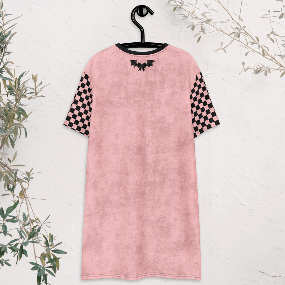 A pink t-shirt dress is shown from the back, hanging on a black hanger against a white textured wall. The dress features black checkerboard sleeves and a small black bow and bat-wing graphic centered near the neckline. Green branches frame the edges of the image.