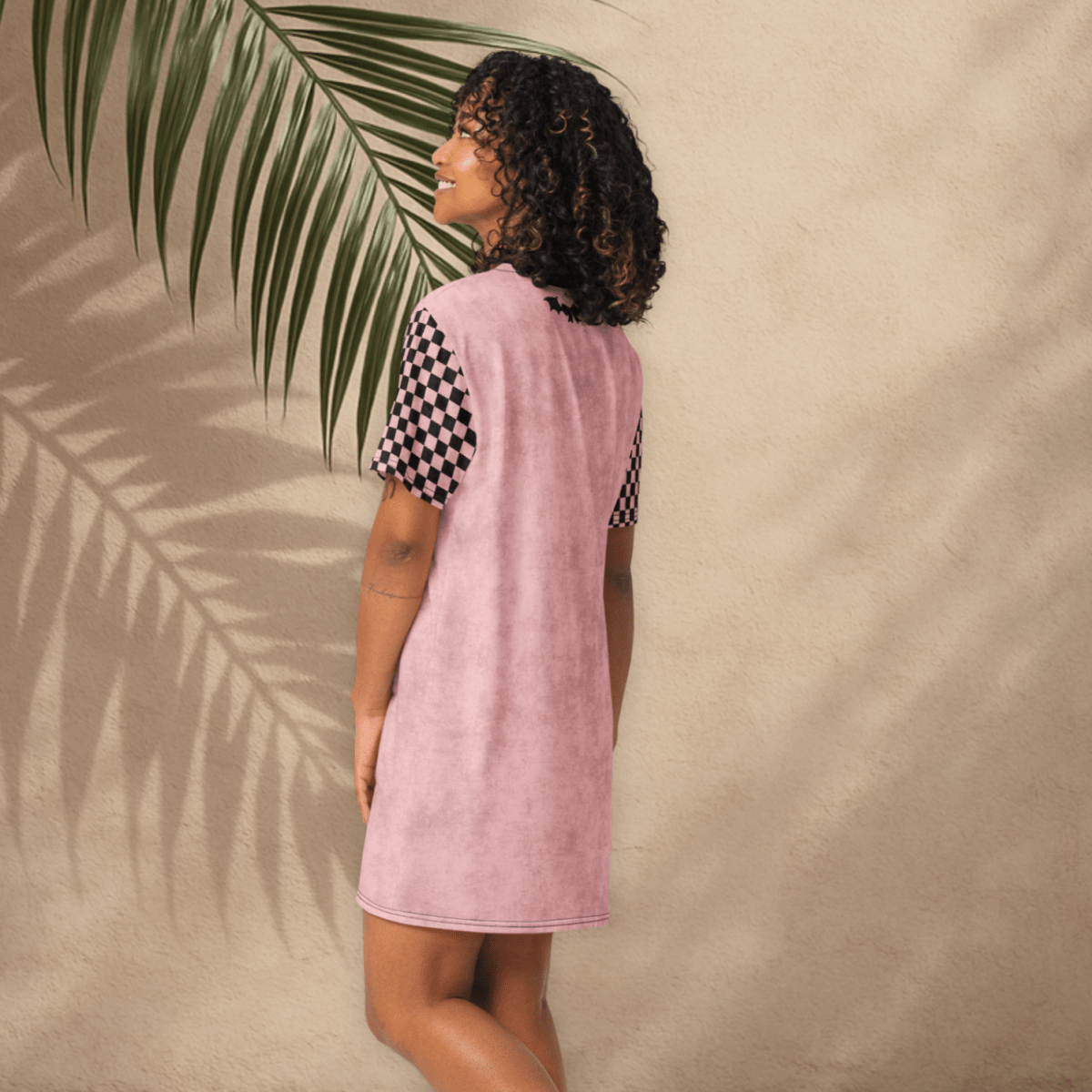 A smiling person with curly dark hair stands with their back partially turned, modeling a pink t-shirt dress with black checkerboard sleeves. The dress features a small black bat-and-bow graphic on the upper back and no visible text on the rear. The setting includes a tan stucco wall and tile floor, with shadows of palm fronds across the background.