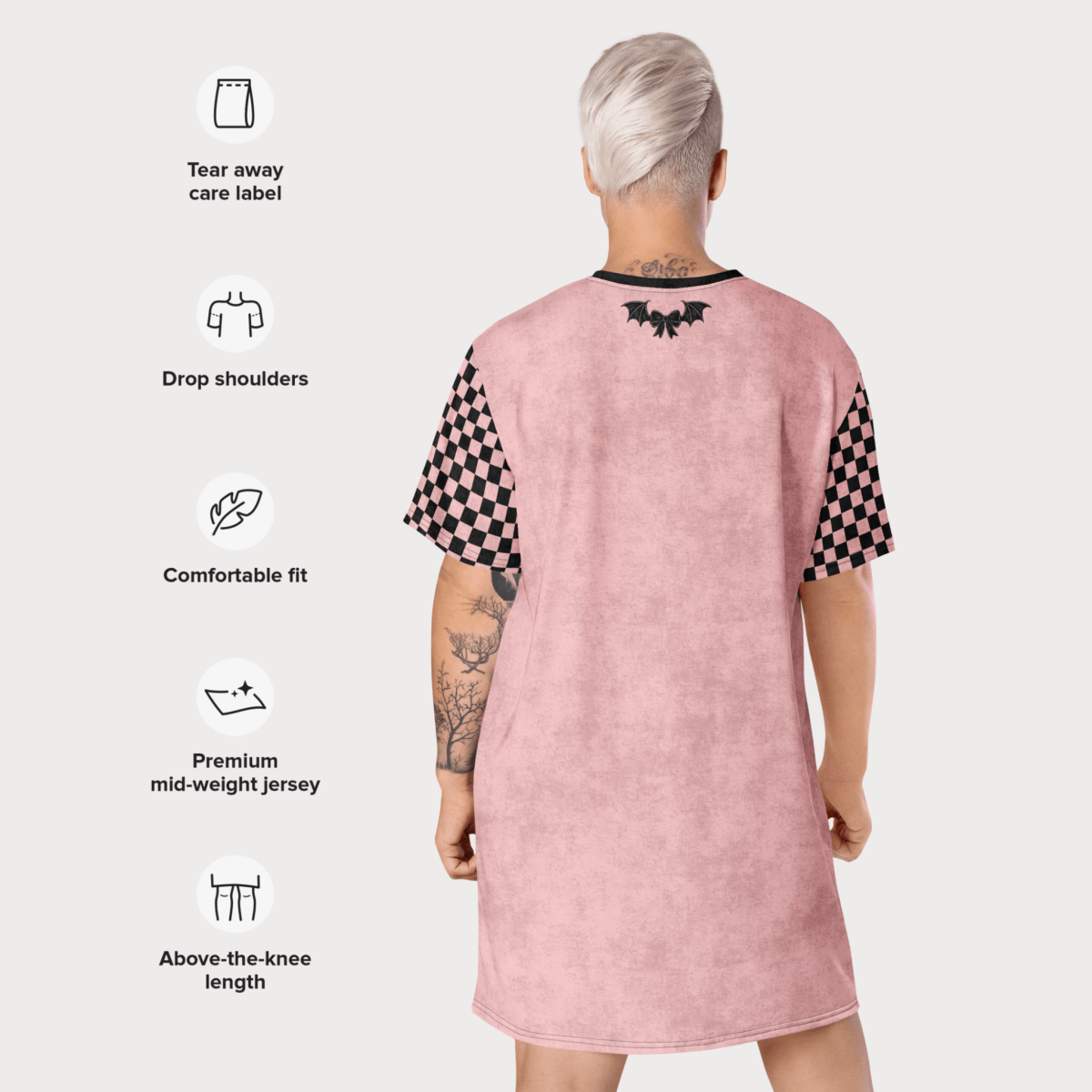 A person with platinum-blonde short hair is shown from behind modeling a pink t-shirt dress. The dress features drop shoulders, black checkerboard sleeves, and a black bat-and-bow graphic at the upper back. Icons and text are overlaid on the left side of the image listing dress features: tear-away care label, drop shoulders, comfortable fit, premium mid-weight jersey, and above-the-knee length.