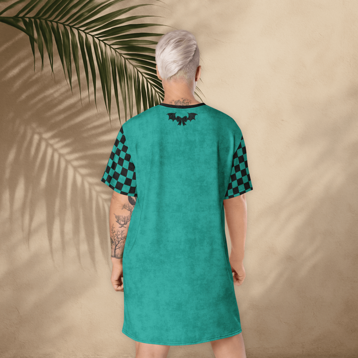 A person with platinum-blonde hair stands with their back to the camera, wearing a teal t-shirt dress. The dress has black checkerboard sleeves and a small black bat-and-bow graphic printed between the shoulder blades. The background is a light tan stucco wall with large palm leaf shadows cast across it. The person’s tattoos are visible on both arms
