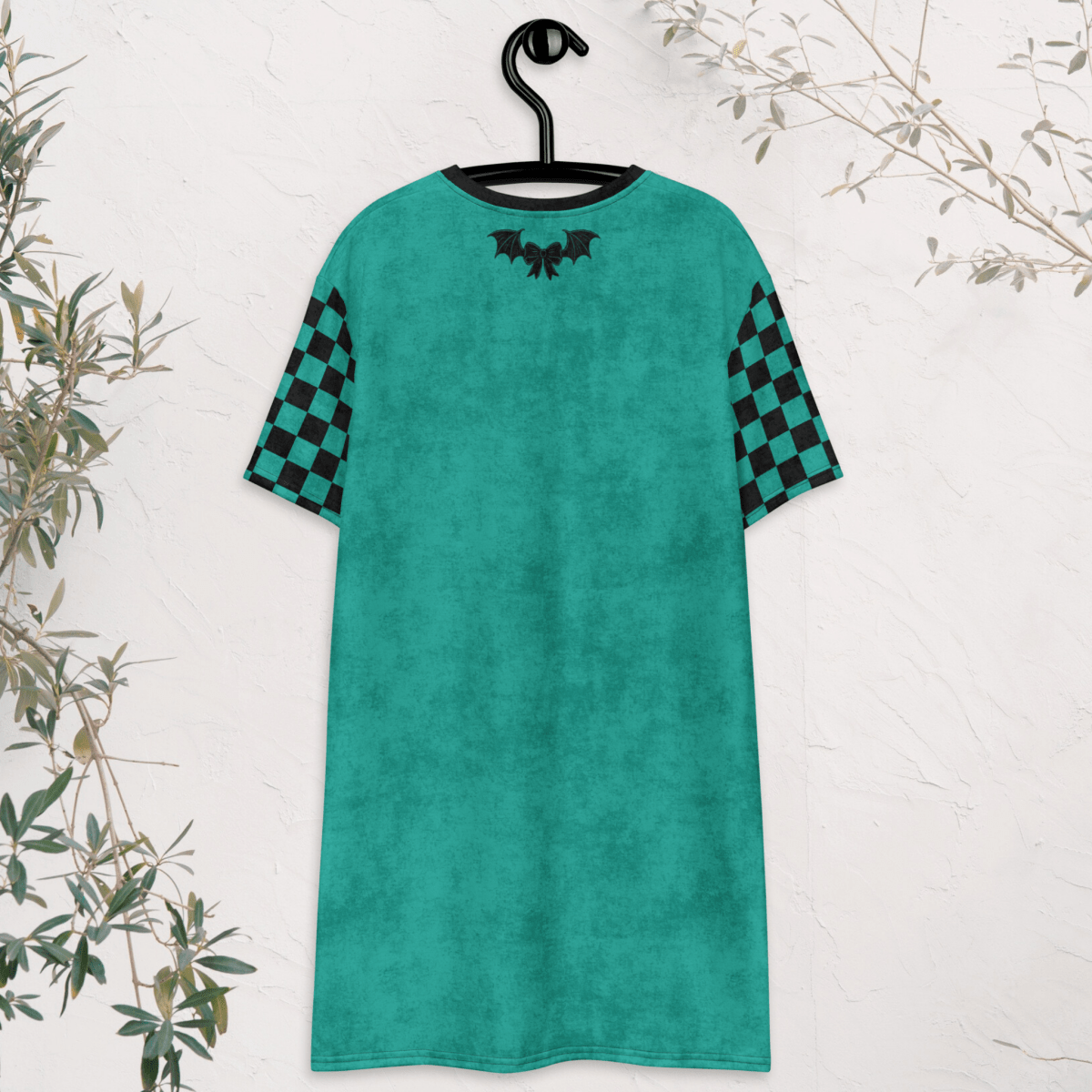 A teal t-shirt dress is shown from the back, hanging on a black hanger against a white textured wall. The dress features black checkerboard sleeves and a small black bow and bat-wing graphic centered near the neckline. Green branches frame the edges of the image.
