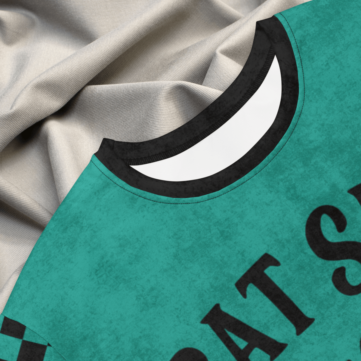 A close-up image shows the collar detail of a teal t-shirt dress laying on gray fabric. The ribbed black collar borders a white interior tag area. The visible portion of the chest includes part of the black printed phrase "BAT SHIT" on the teal fabric with faint textured patterning.