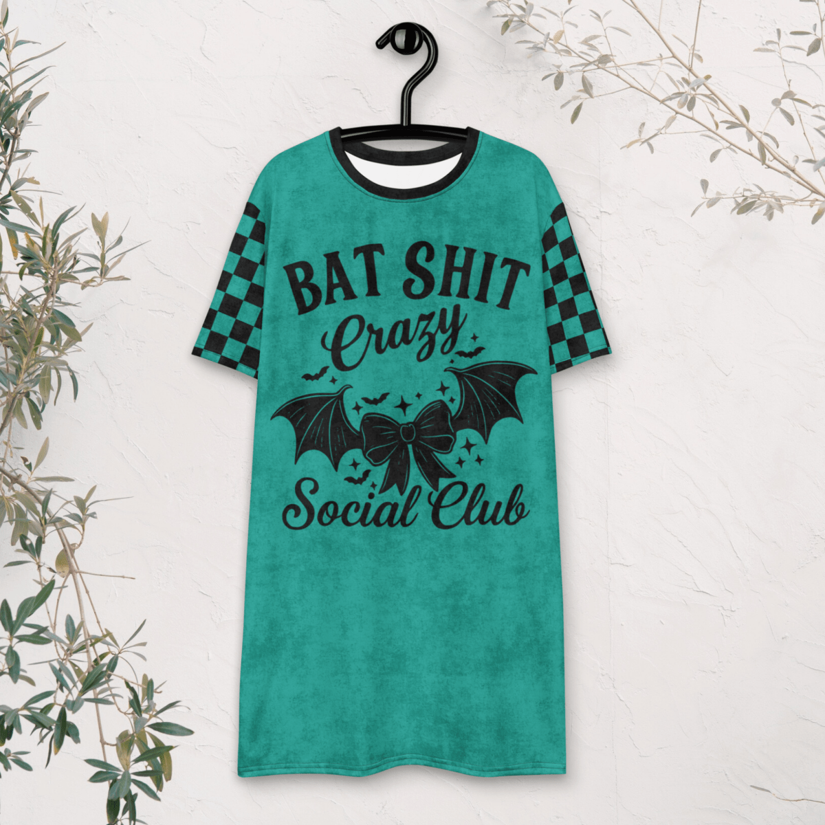A teal t-shirt dress is displayed on a black hanger against a white textured wall. The dress features black checkerboard short sleeves and a large front graphic in black that says "BAT SHIT Crazy Social Club," surrounded by bats, stars, and a central bow. Some foliage partially frames the edge of the photo.