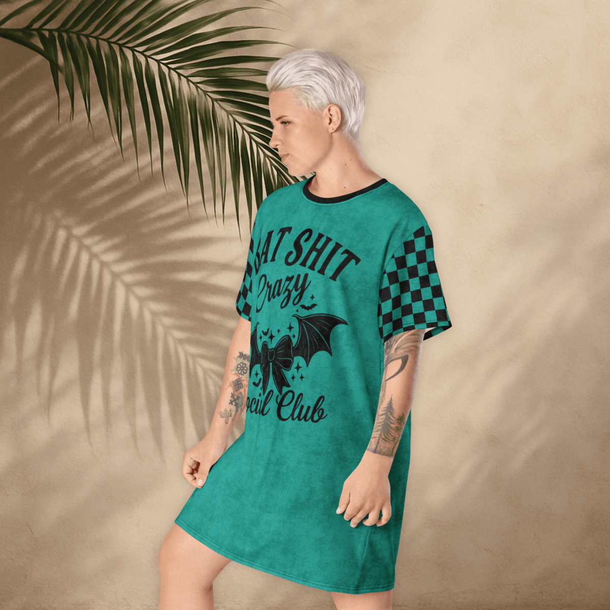 A person with platinum-blonde hair stands in a teal t-shirt dress with a moody expression and one foot slightly raised. The dress features black checkerboard sleeves and a large black front print reading "BAT SHIT Crazy Social Club" with a decorative bow and bat wings. The tan background includes soft palm shadows.