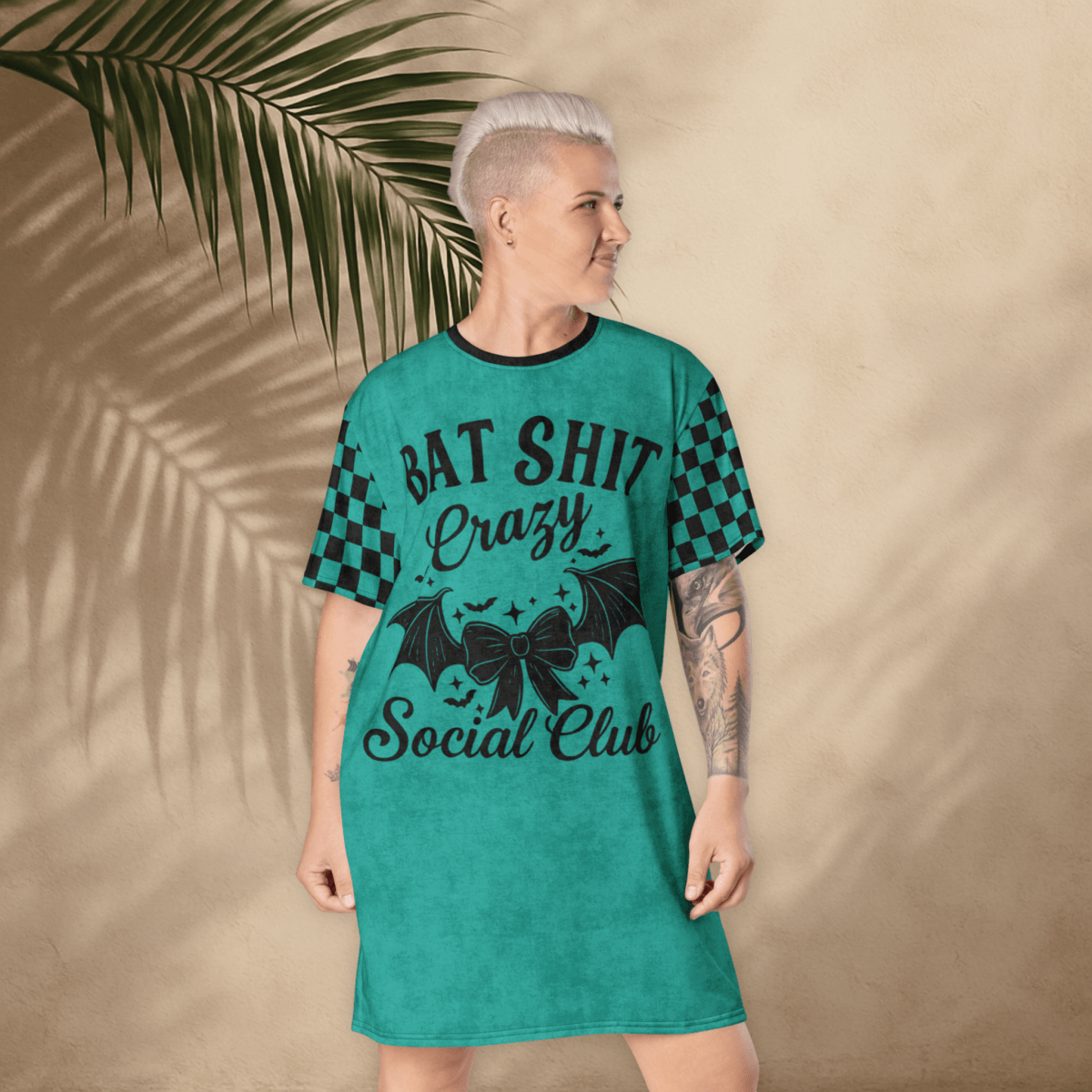 A person with platinum-blonde hair stands with their back to the camera, wearing a teal t-shirt dress. The dress has black checkerboard sleeves and a small black bat-and-bow graphic printed between the shoulder blades. The background is a light tan stucco wall with large palm leaf shadows cast across it. The person’s tattoos are visible on both arms
