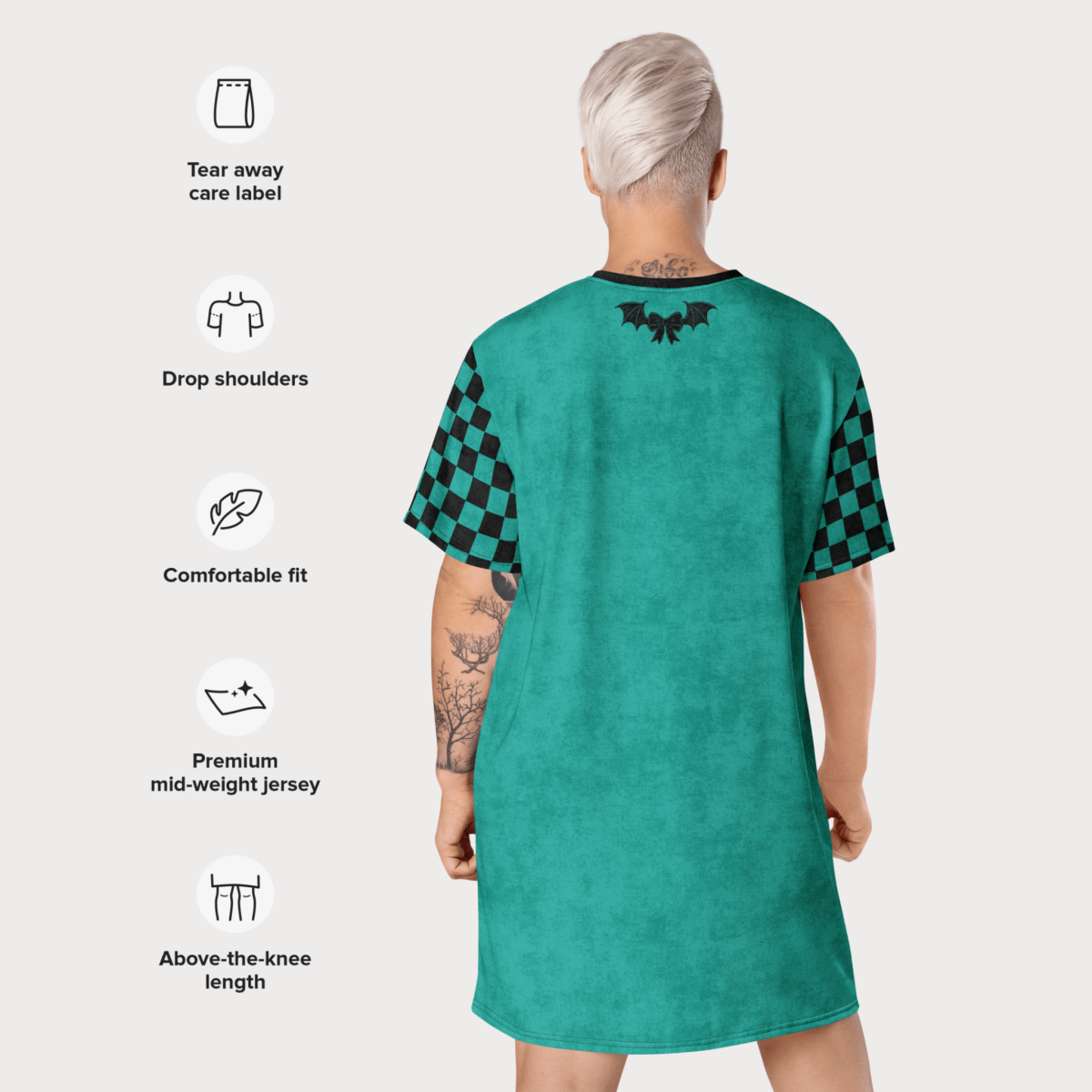 A person with platinum-blonde short hair is shown from behind modeling a teal t-shirt dress. The dress features drop shoulders, black checkerboard sleeves, and a black bat-and-bow graphic at the upper back. Icons and text are overlaid on the left side of the image listing dress features: tear-away care label, drop shoulders, comfortable fit, premium mid-weight jersey, and above-the-knee length.