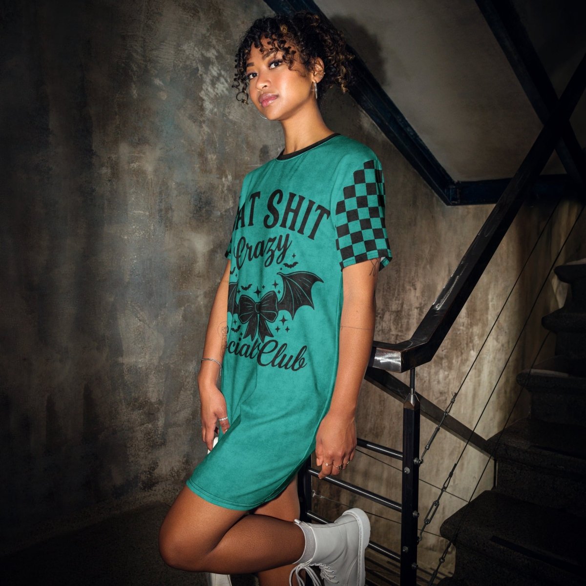 A young woman with curly hair stands on an industrial-style staircase in front of a weathered concrete wall. She wears a teal t-shirt dress with black checkerboard sleeves and a large black graphic on the front that says "BAT SHIT Crazy Social Club." The design includes a bow with bat wings, small stars, and bold gothic text. She pairs the dress with white lace-up boots and gazes confidently at the camera.