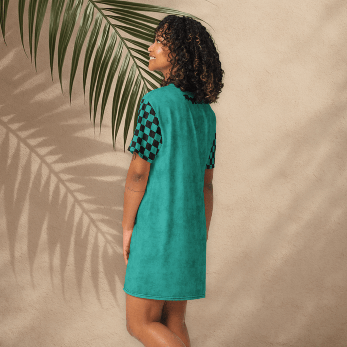A smiling person with curly dark hair stands with their back partially turned, modeling a teal t-shirt dress with black checkerboard sleeves. The dress features a small black bat-and-bow graphic on the upper back and no visible text on the rear. The setting includes a tan stucco wall and tile floor, with shadows of palm fronds across the background.