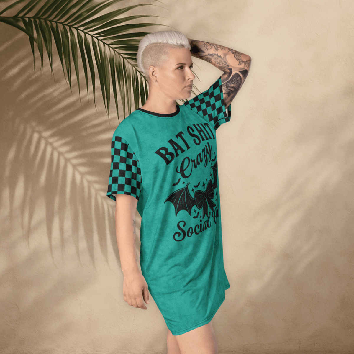 A person with platinum-blonde hair and arm tattoos poses in a teal t-shirt dress against a tan wall with palm leaf shadows. The dress features black checkerboard sleeves and a bold black graphic that reads "BAT SHIT Crazy Social Club" with a bat-winged bow and stars. The person stands with one hand on their head, tilting slightly forward.