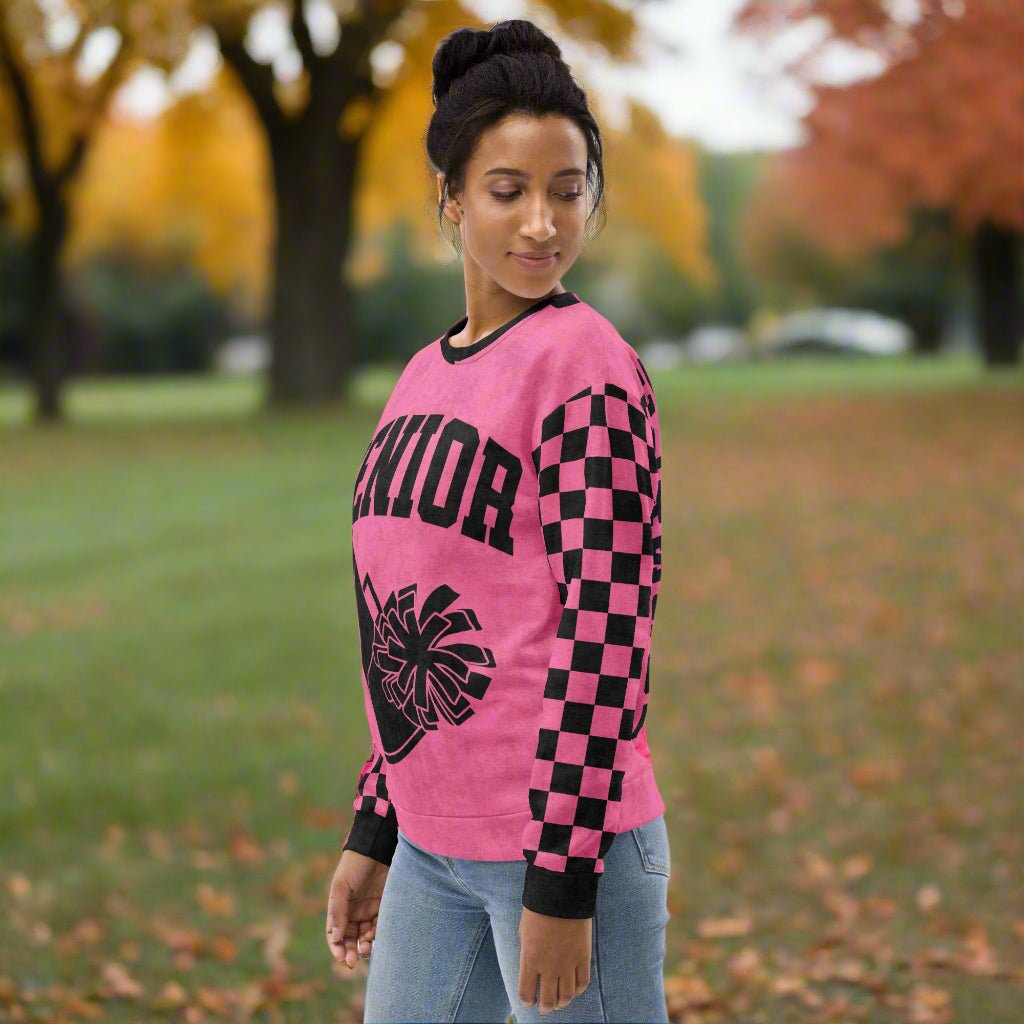 Person wearing a pink long-sleeve sweatshirt shirt with black checkered sleeves and a senior cheerleading design outdoors