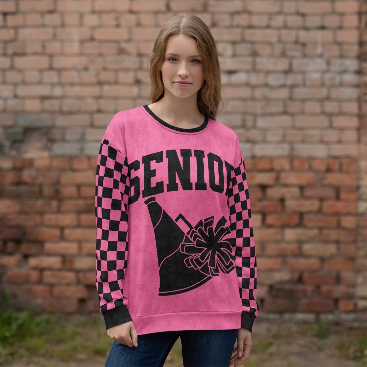 Person wearing a pink sweater with black checkered pattern and 'SENIOR' text with pom pom and megaphone against a brick wall.
