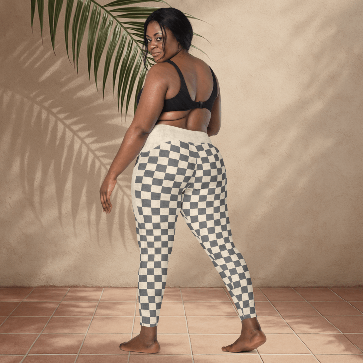 Woman wearing checkered pants standing against a beige wall with a plant leaf.