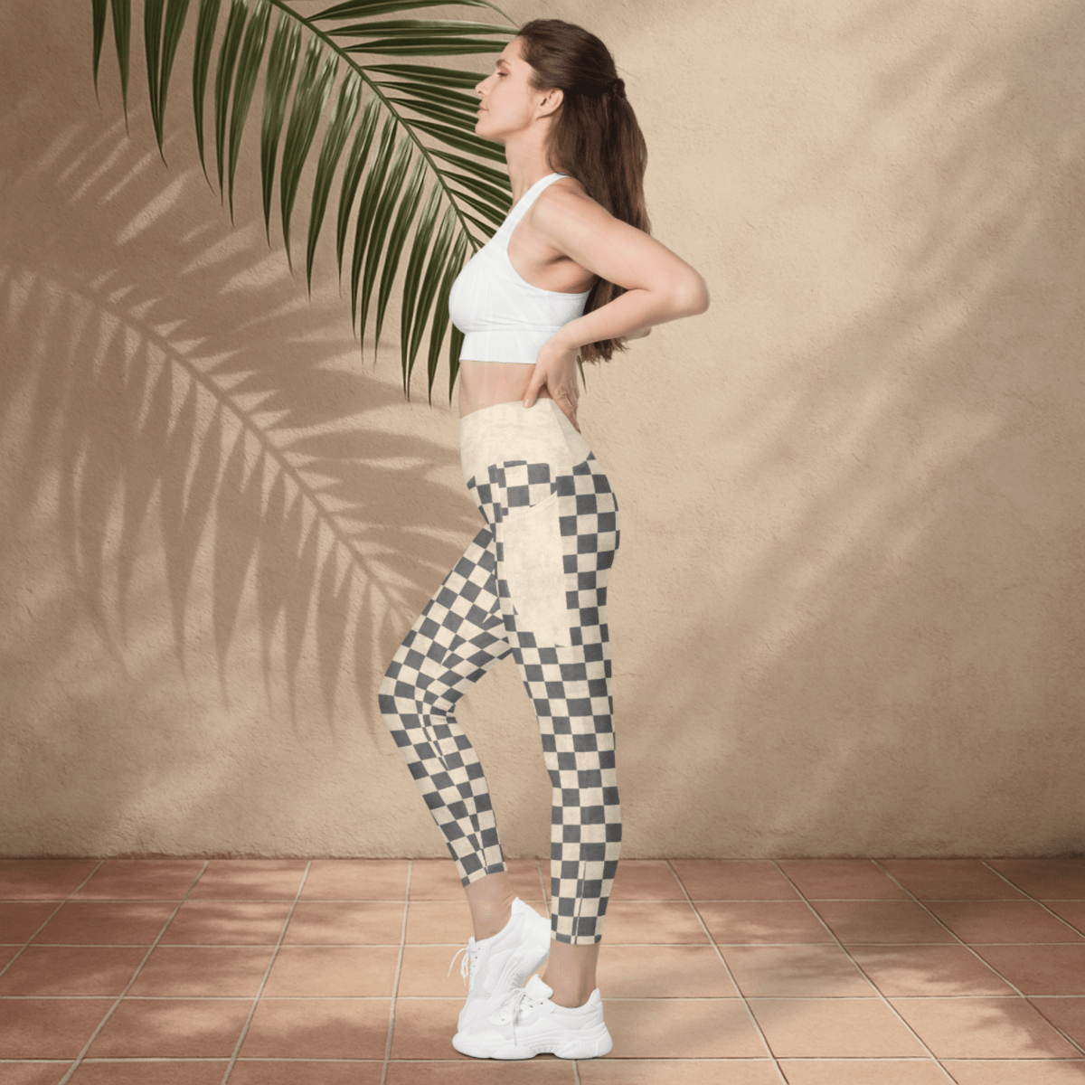 Woman wearing a white sports bra and checkered leggings standing against a beige wall with a palm leaf.