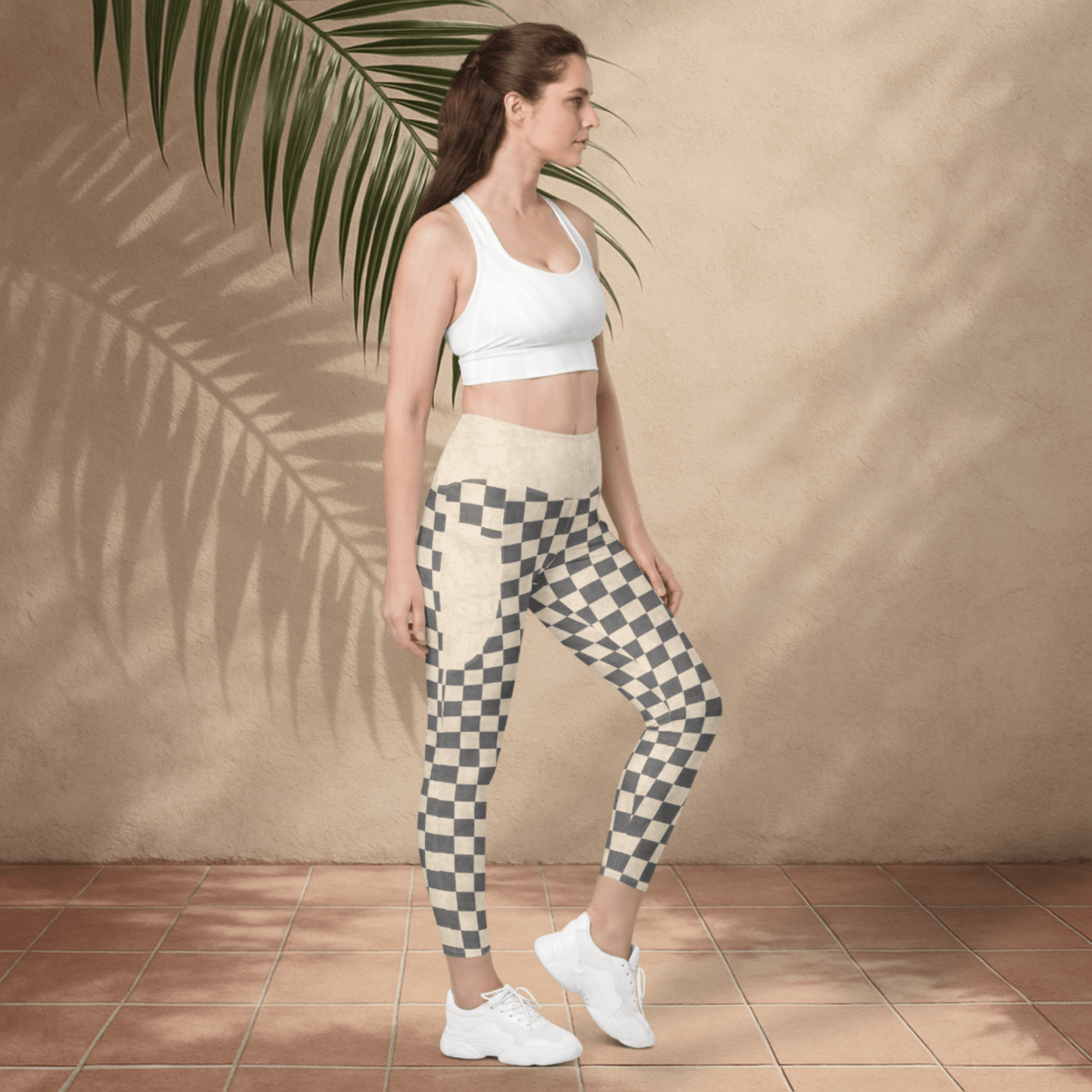 Woman wearing a white sports bra and checkered leggings standing against a beige wall with a palm leaf.