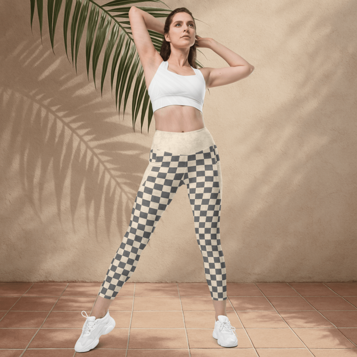 Woman wearing a white sports bra and checkered leggings standing against a beige wall with a palm leaf.