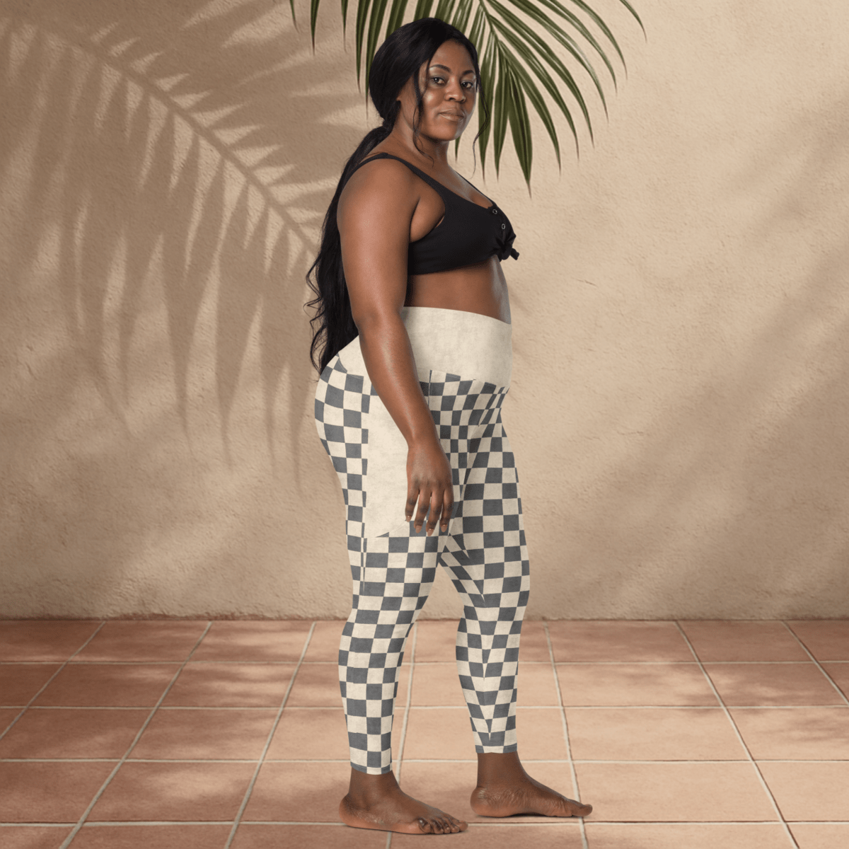 Woman wearing a black top and checkered pants standing against a beige wall with a plant.