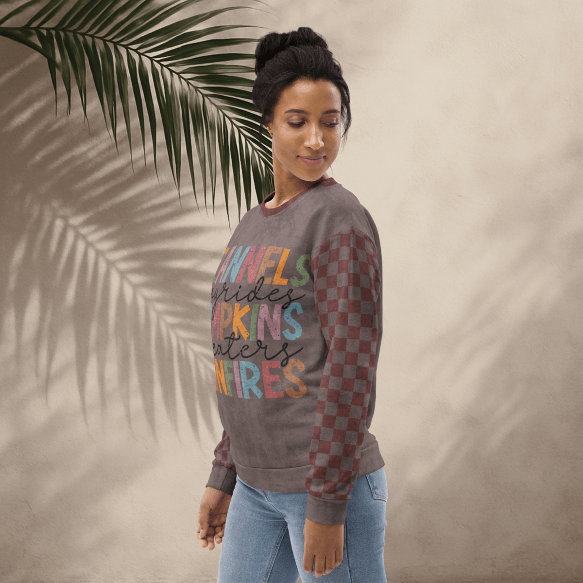Woman wearing an espresso and burgundy sweater with colorful text design, standing against a beige wall with a palm leaf.