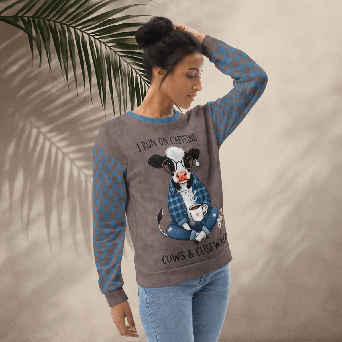 Woman wearing Espresso and denim Sweatshirt with a cow graphic and text, standing against a beige wall with a palm leaf.