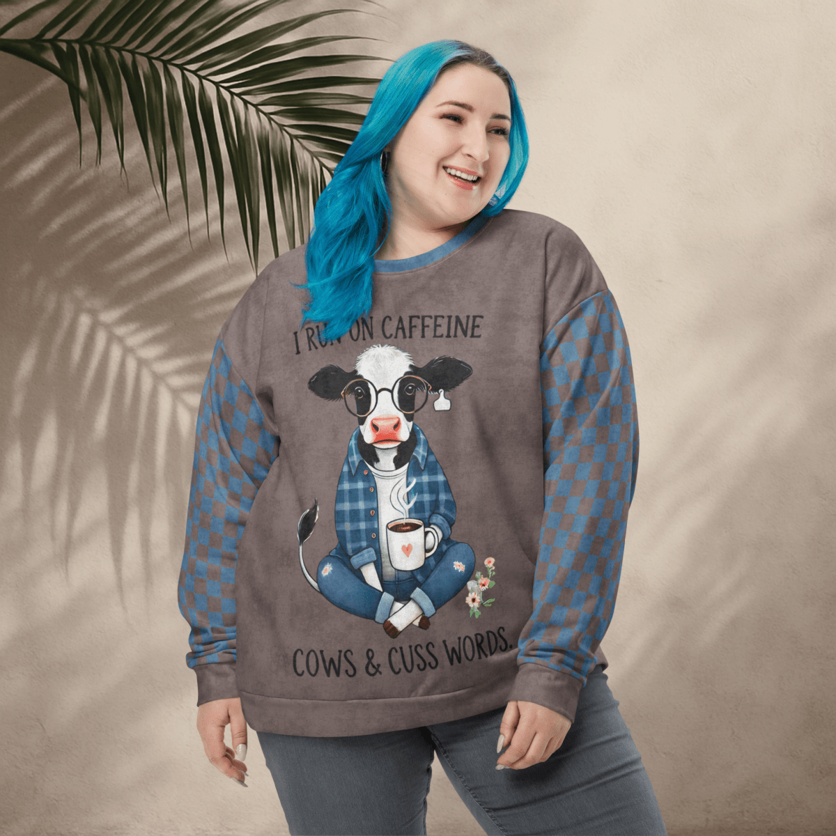 Person wearing Espresso and denim Sweatshirt with a cow graphic and text, standing against a beige wall with a leafy branch.