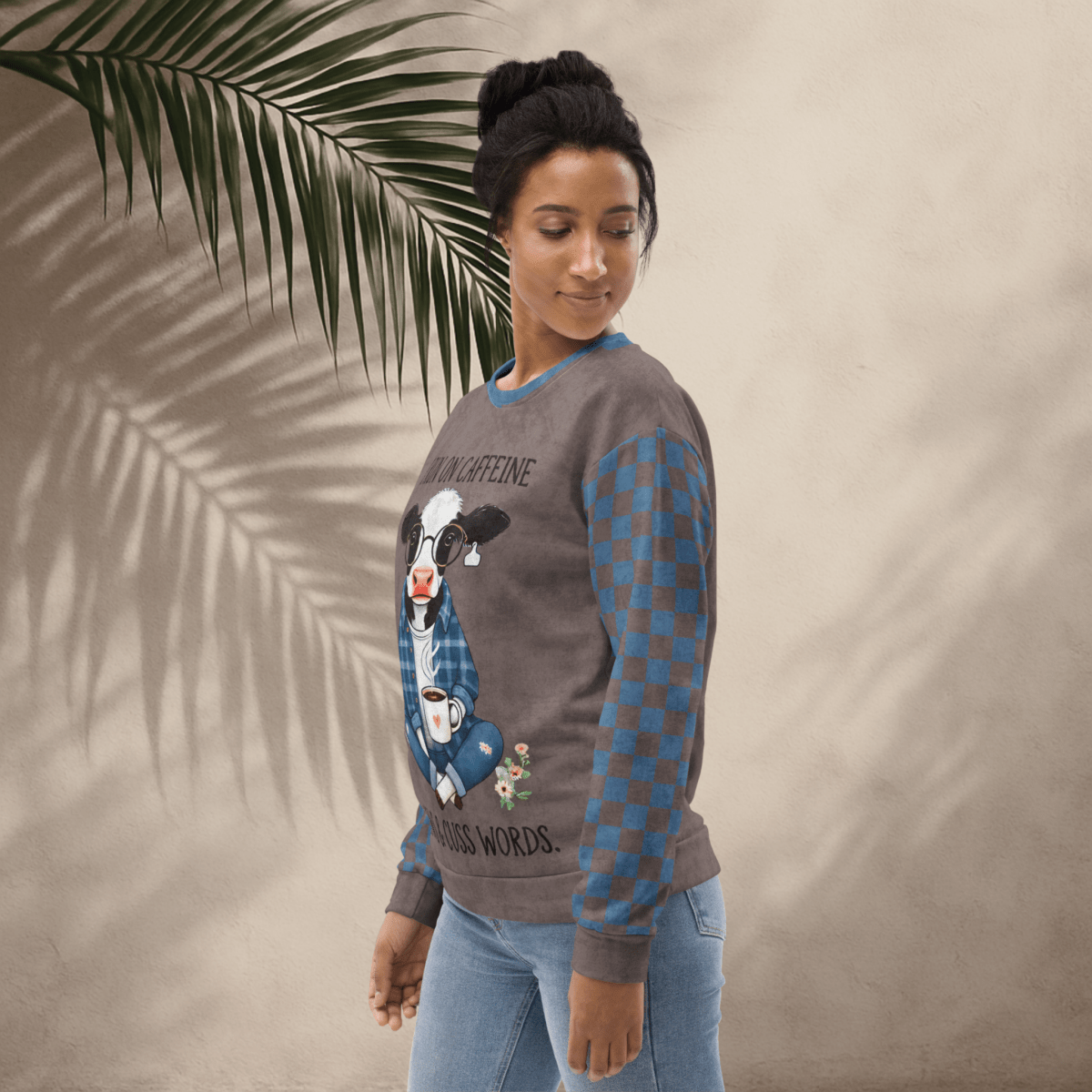 Woman wearing Espresso and denim Sweatshirt with a cow design and text, standing against a beige wall with a palm leaf.