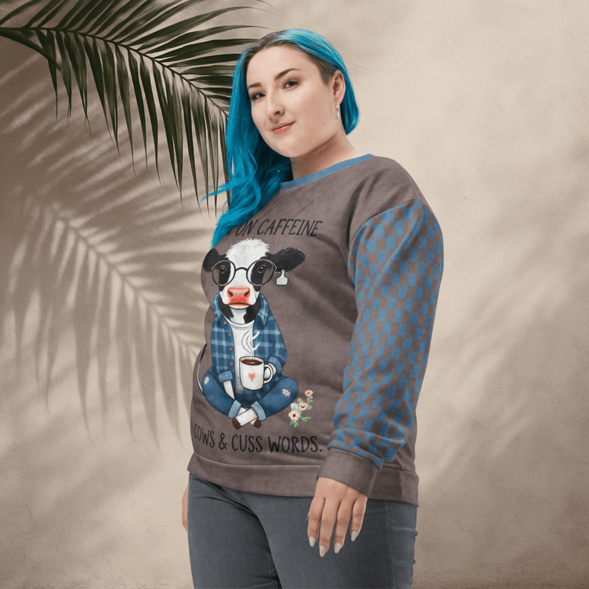 Person wearing Espresso and denim Sweatshirt with a cow design and text, standing against a beige wall with a leafy branch.