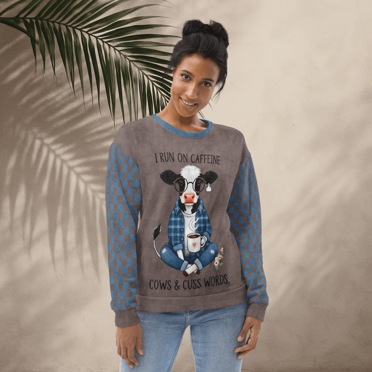 Woman wearing Espresso and denim Sweatshirt with a cow graphic and text, standing against a beige wall with a leafy branch.