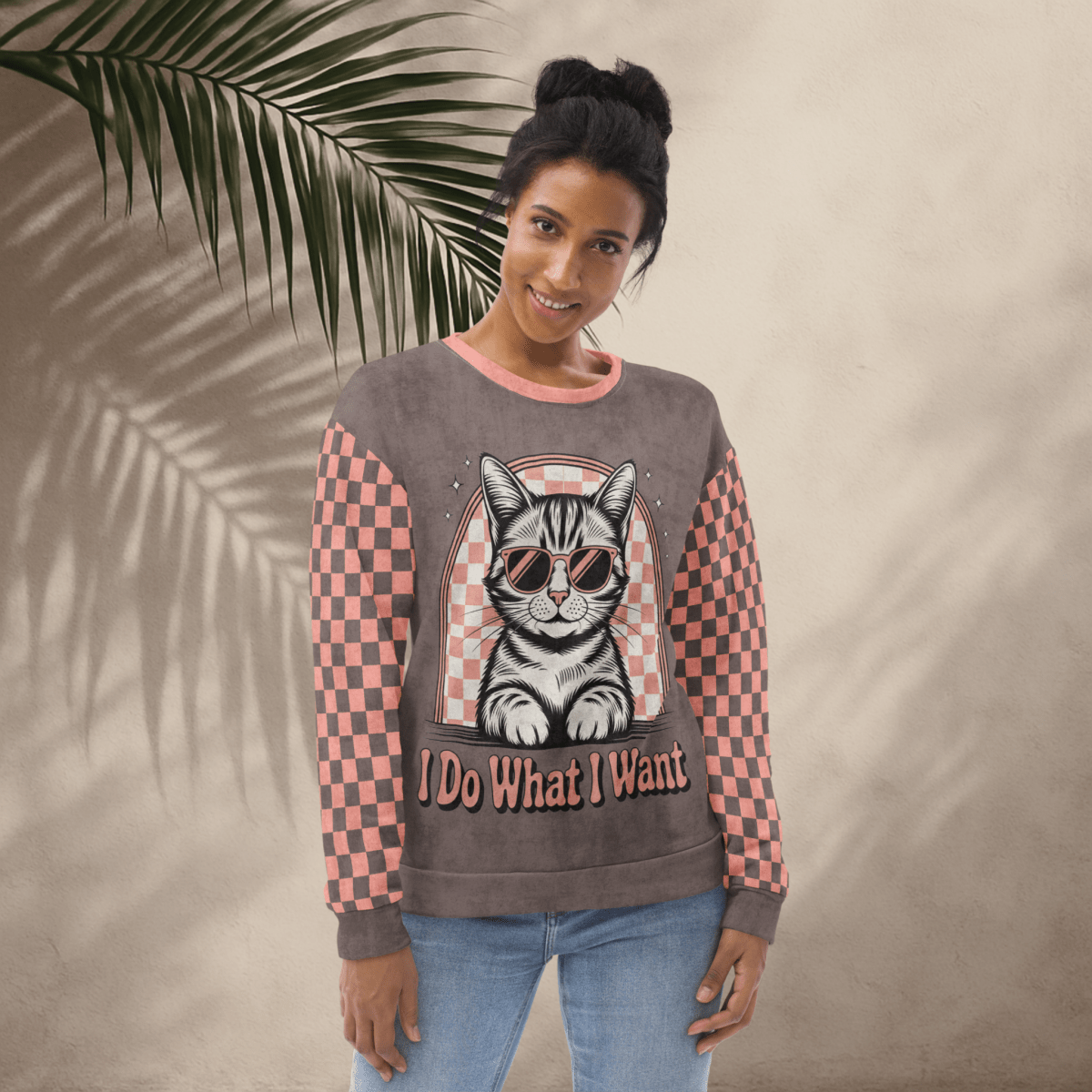 model wearing Espresso sweatshirt with a sunglasses-wearing tabby cat and “I Do What I Want” text; sleeves feature pink-and-espresso checkerboard pattern with pink collar and cuffs.
