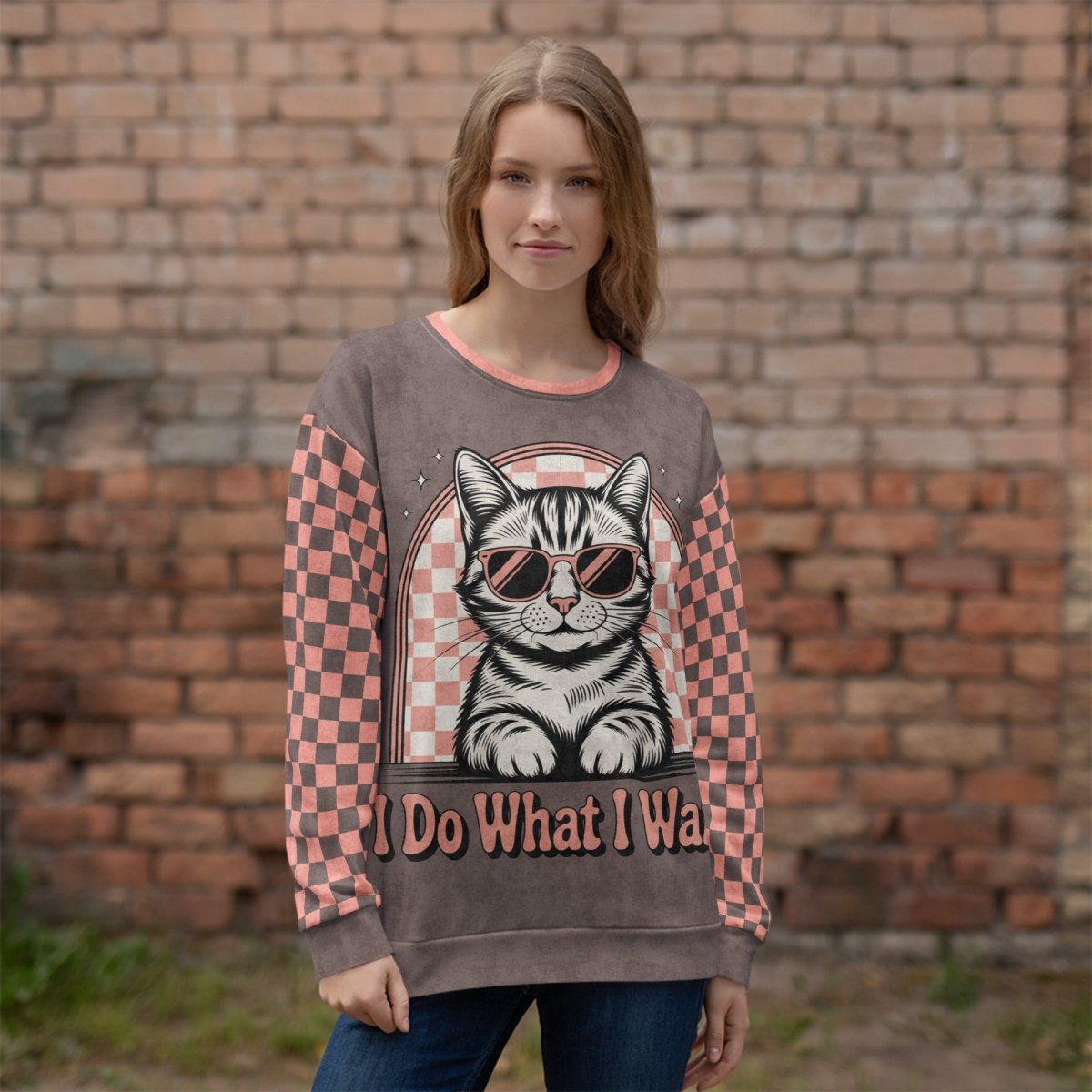 model wearing an Espresso sweatshirt with a sunglasses-wearing tabby cat and “I Do What I Want” text; sleeves feature pink-and-espresso checkerboard pattern with pink collar and cuffs.