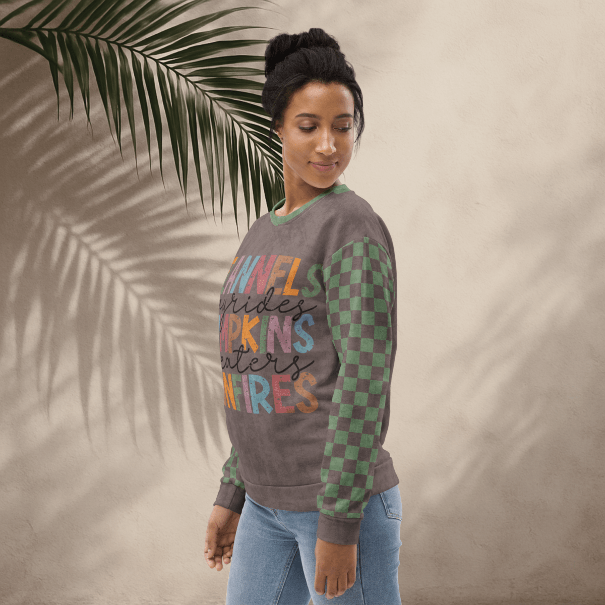Woman wearing an Espresso and sage green sweater with text, standing against a beige wall with a palm leaf.