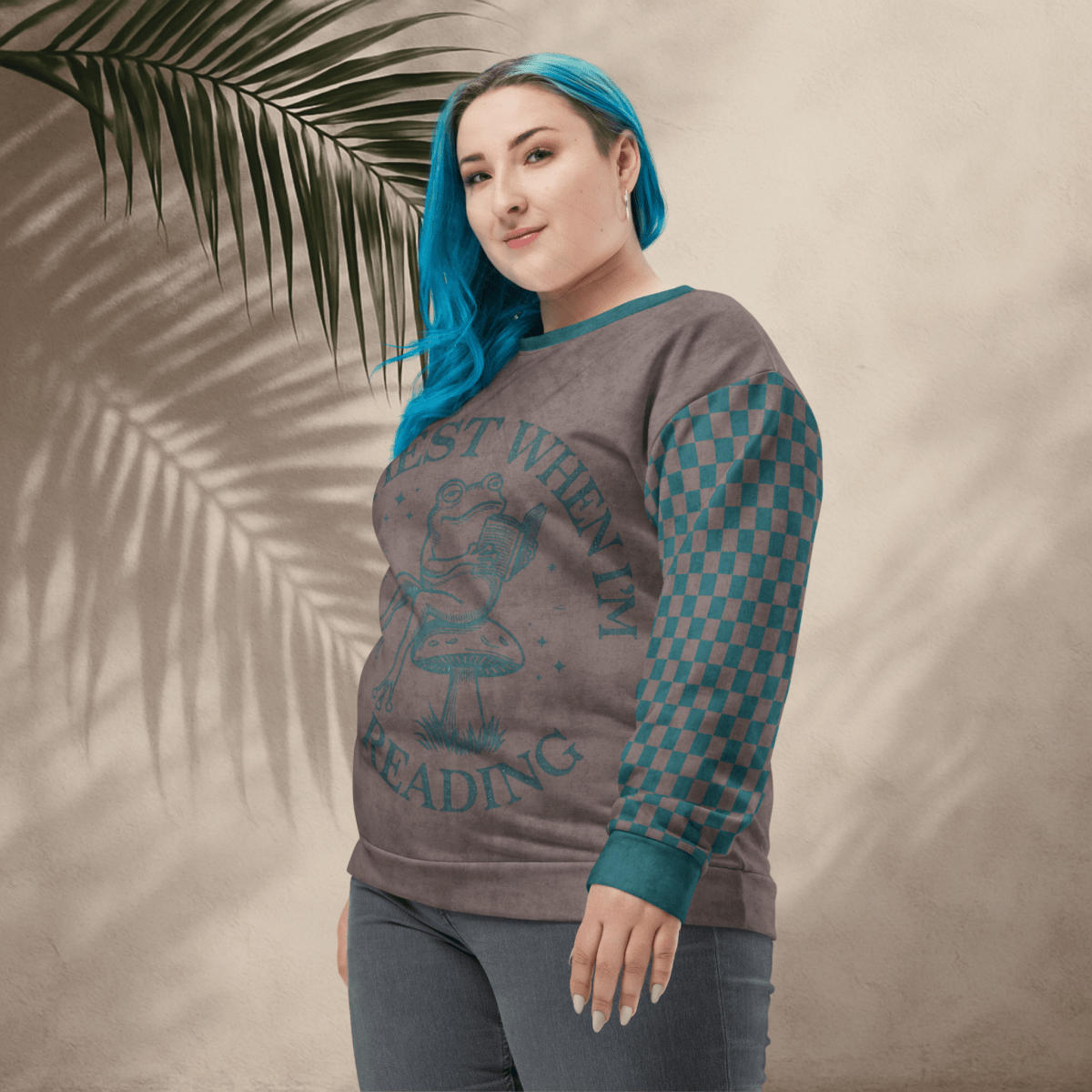 Person wearing an espresso and teal sweatshirt with a Frog reading with "Happiest When I'm Reading" and checkered sleeves against a beige background