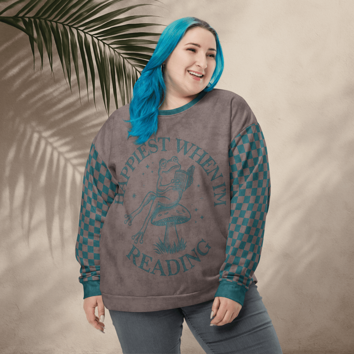 Person wearing an espresso and teal sweatshirt with a Frog reading with "Happiest When I'm Reading" , standing against a beige wall with a leafy branch.