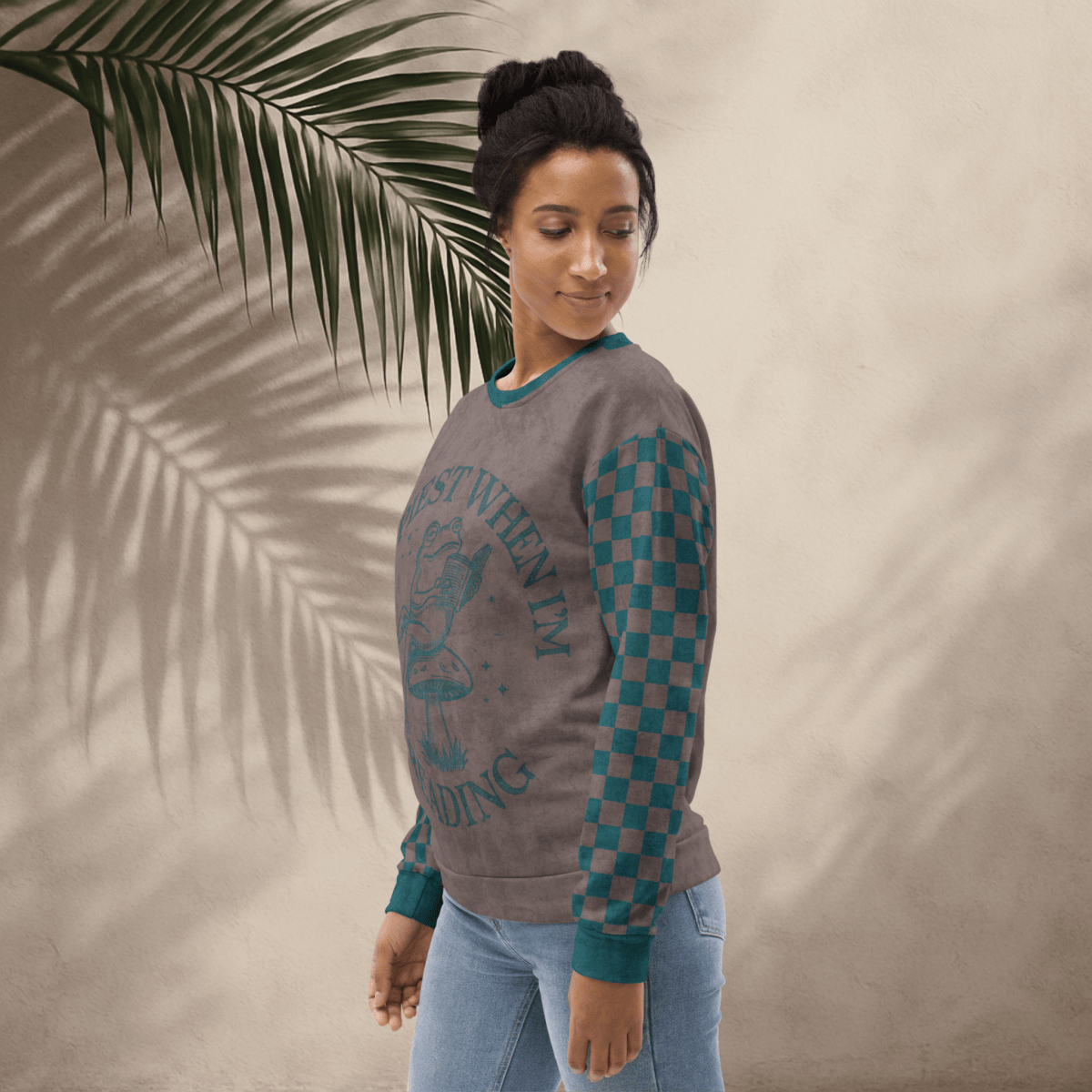 Woman wearing an espresso and teal sweatshirt with a Frog reading with "Happiest When I'm Reading" design and checkered sleeves against a beige background