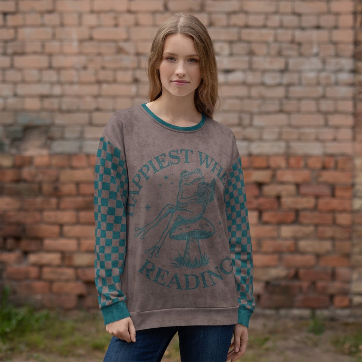 Woman wearing an espresso and teal sweater with a Frog reading with "Happiest When I'm Reading" text design in front of a brick wall
