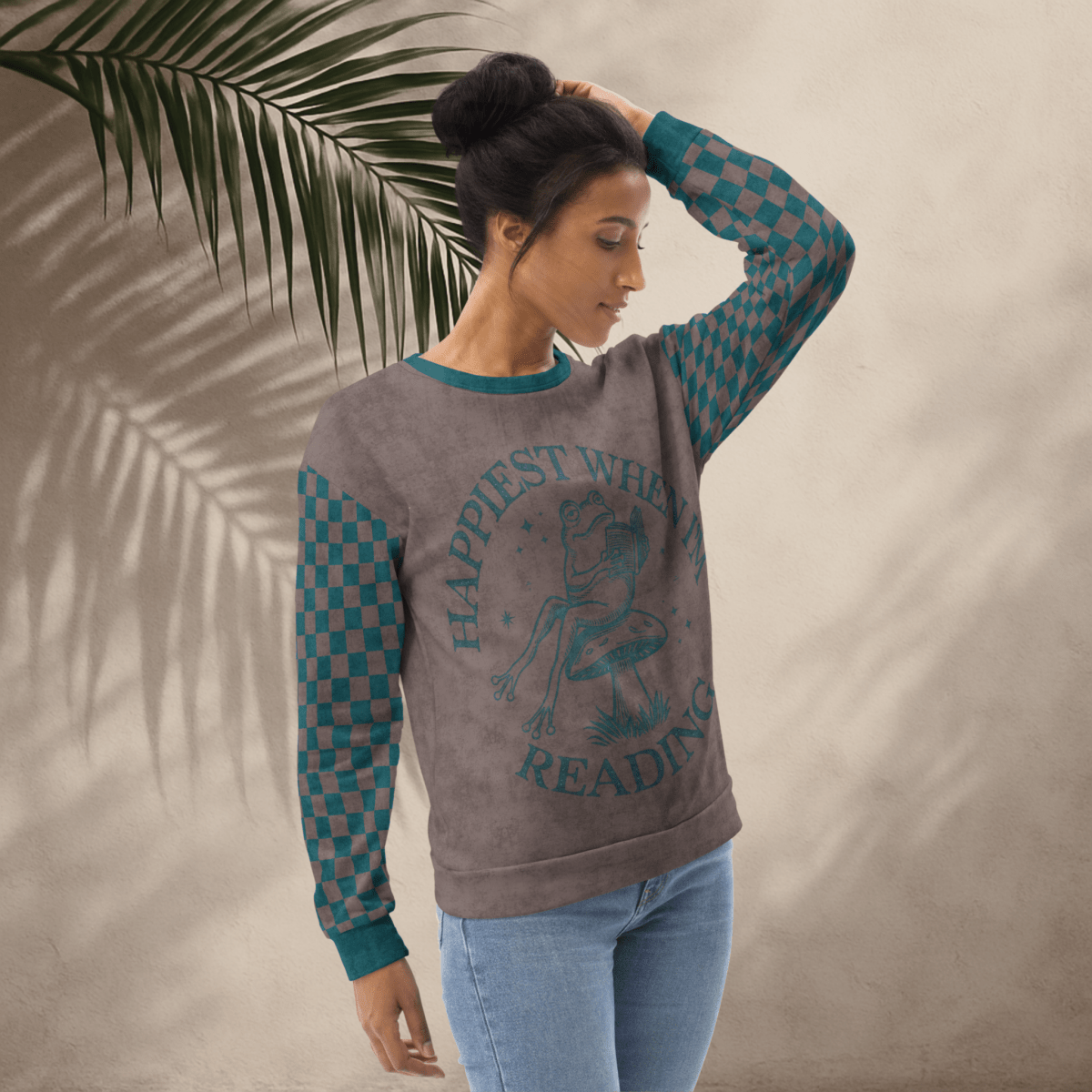 Woman wearing an espresso and teal sweatshirt with a Frog reading with "Happiest When I'm Reading" design and checkered sleeves against a beige background