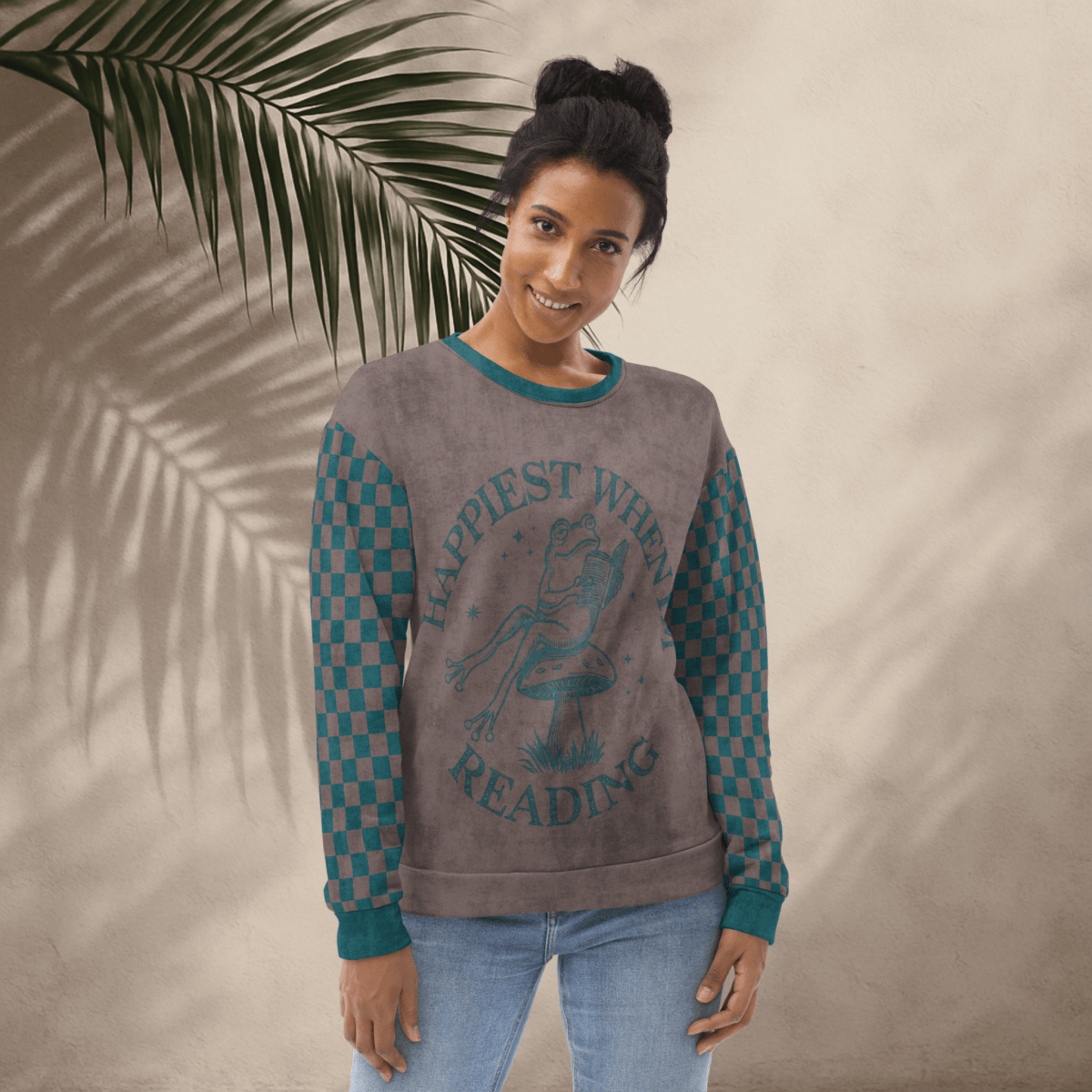 Woman wearing an espresso and teal sweatshirt with a Frog reading with "Happiest When I'm Reading" and checkered sleeves, standing against a beige wall with a palm leaf.