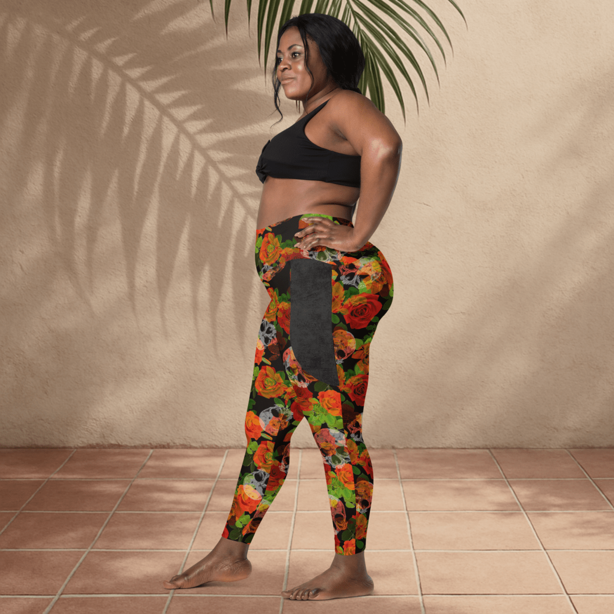 Woman wearing a black sports bra and colorful floral leggings with skulls against a beige wall.
