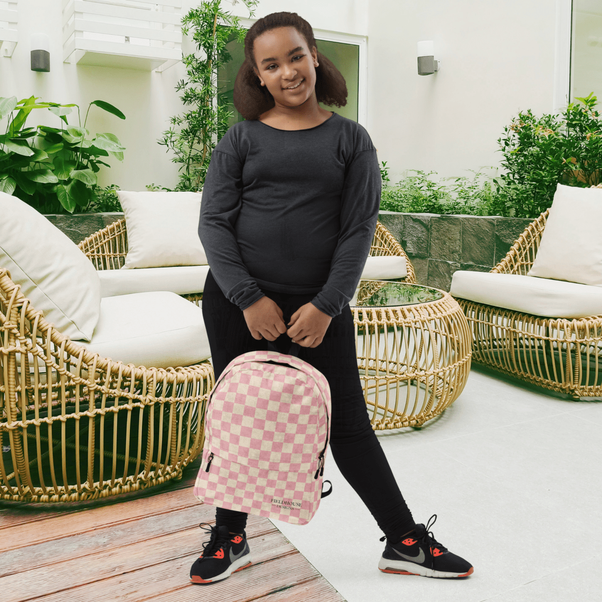 Person holding a pink checkered backpack in an outdoor setting with wicker furniture and plants.